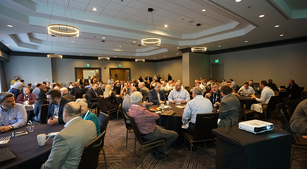 Attendees at the CONSULT Conference engage in networking and discussions in a modern conference room with round tables, a projector, and contemporary lighting.