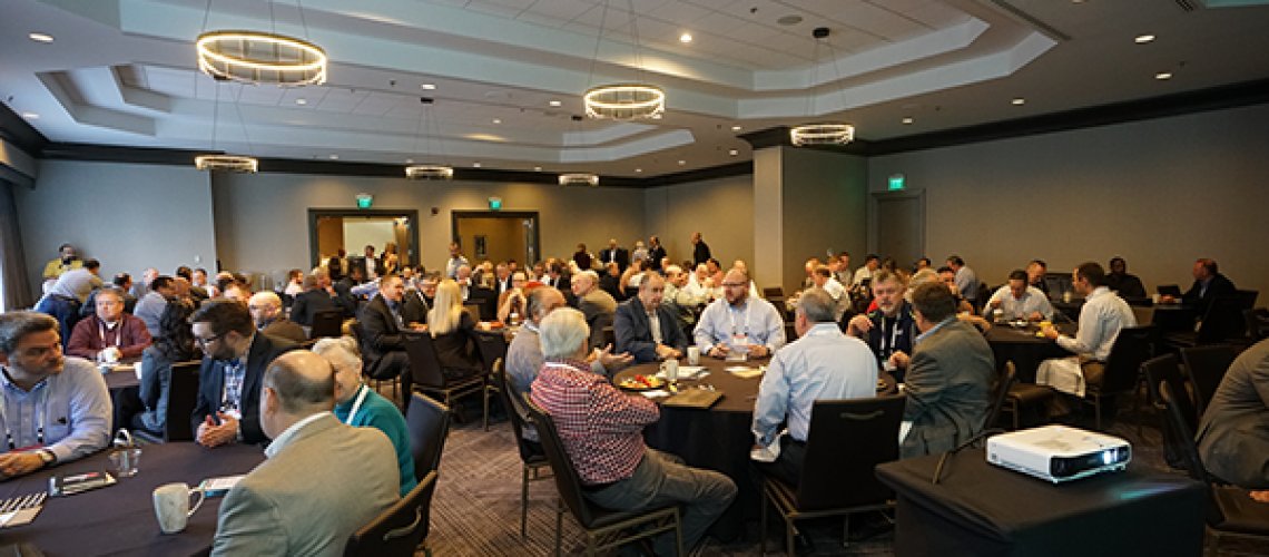 Attendees at the CONSULT Conference engage in networking and discussions in a modern conference room with round tables, a projector, and contemporary lighting.